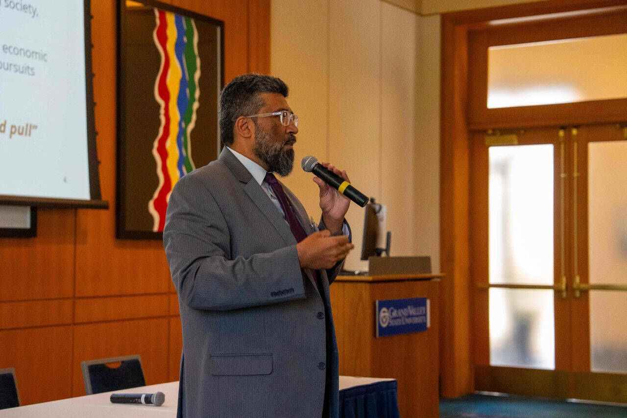 Samir Iqbal, associate dean of research in the College of Computing, speaks into a microphone during his keynote “Tech Transfer from Lab to Market” at Tech Week Grand Rapids. A projection screen with presentation slides is visible beside him.